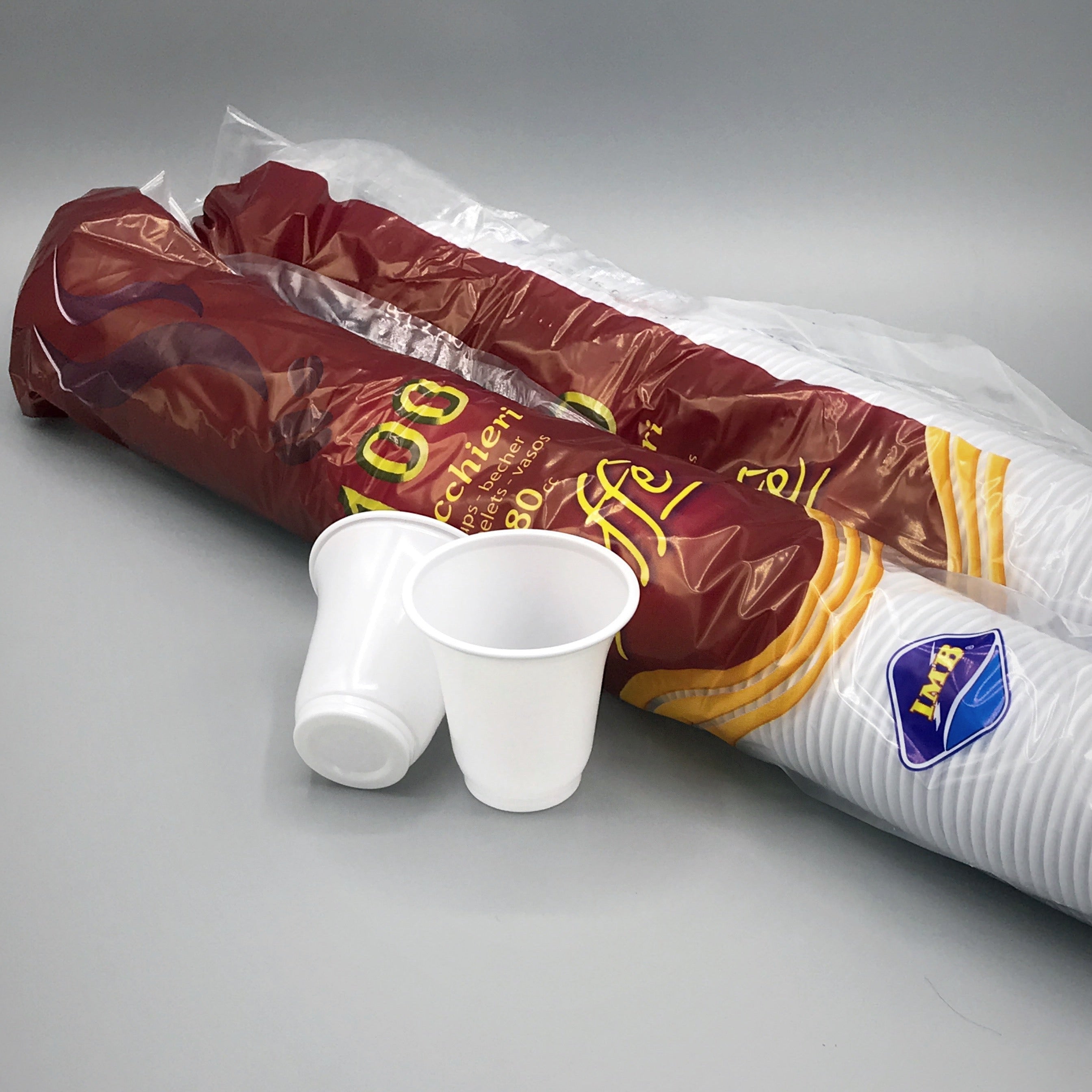A bulk pack of disposable plastic cups, including two loose white cups in the foreground and a large roll of white cups wrapped in a clear sleeve and dark red paper band with gold lettering, set against a plain gray background.