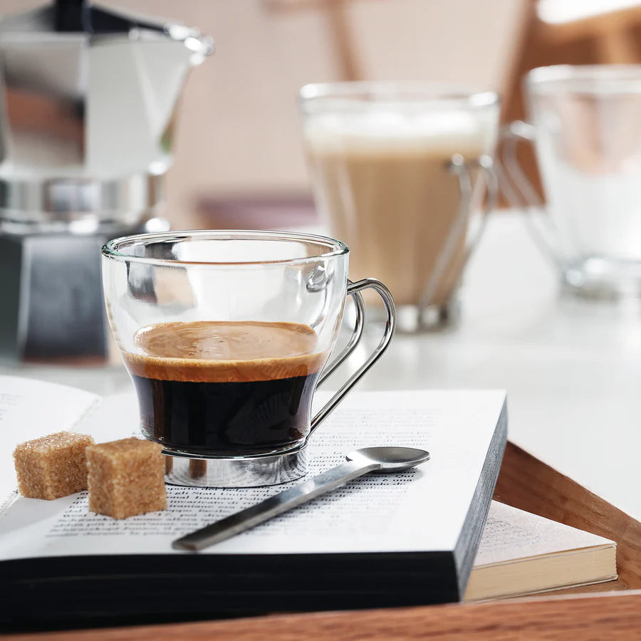 A clear glass mug filled with espresso sits on an open book, with brown sugar cubes and a spoon nearby. A moka pot and other cups are blurred in the background on a tabletop.